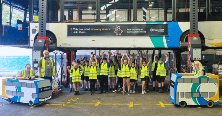 The class of children in specially designed high-vis under a bus on a ramp at Stagecoach East's Peterborough Depot