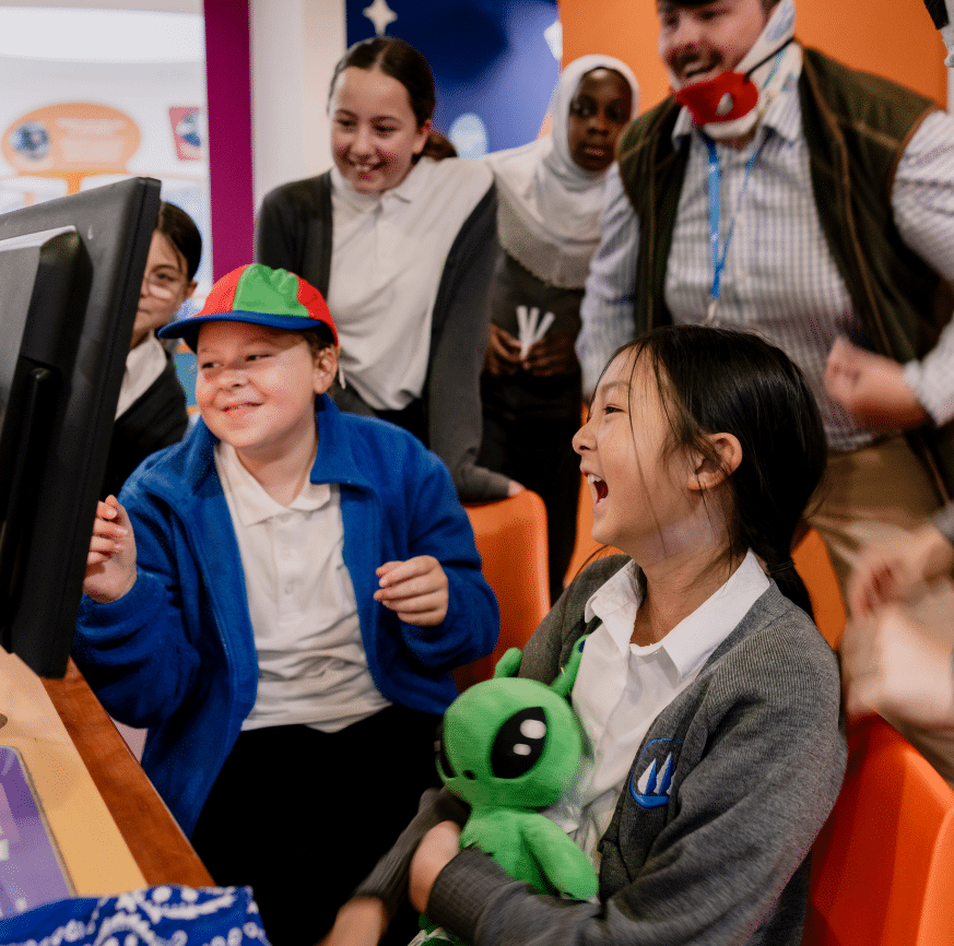 Children - one with an alien in her lap and another wearing a coloured cap, surrounded by classmates gather around a computer screen laughing