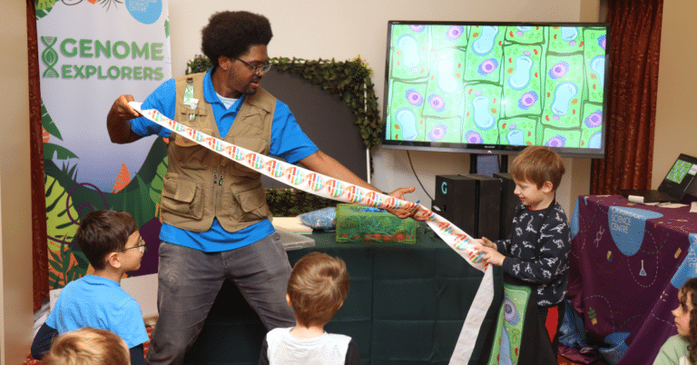 A child helps a science communicator unravel a long strip of paper, representing a chain of DNA