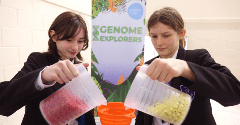 Two school girls pour yellow and red beads into a funnel in front of a roll up banner tilted Genome Explorers